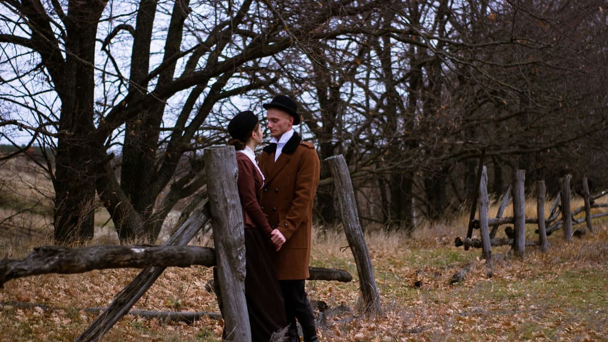 A couple standing by a rustic fence in a serene autumn landscape, sharing a tender moment.