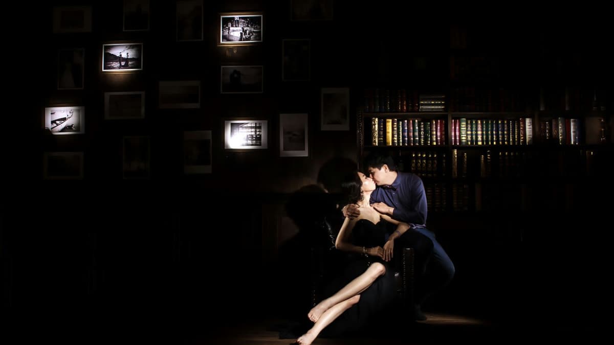 A romantic moment captured in a dark library with a couple kissing, surrounded by illuminated books and photos.