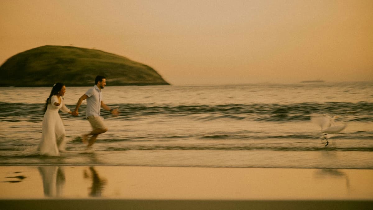 A couple runs joyfully on a beach at sunset with a scenic view of Rio de Janeiro.