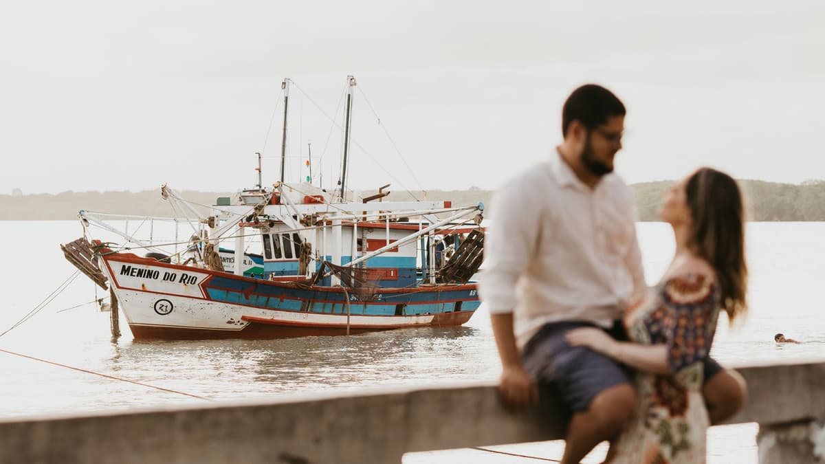 A loving couple sits by a calm lake with a fishing boat in the background, evoking romance and tranquility.