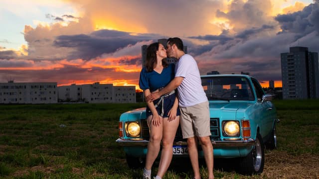 A couple embraces near a vintage blue car at sunset with a vibrant sky backdrop.