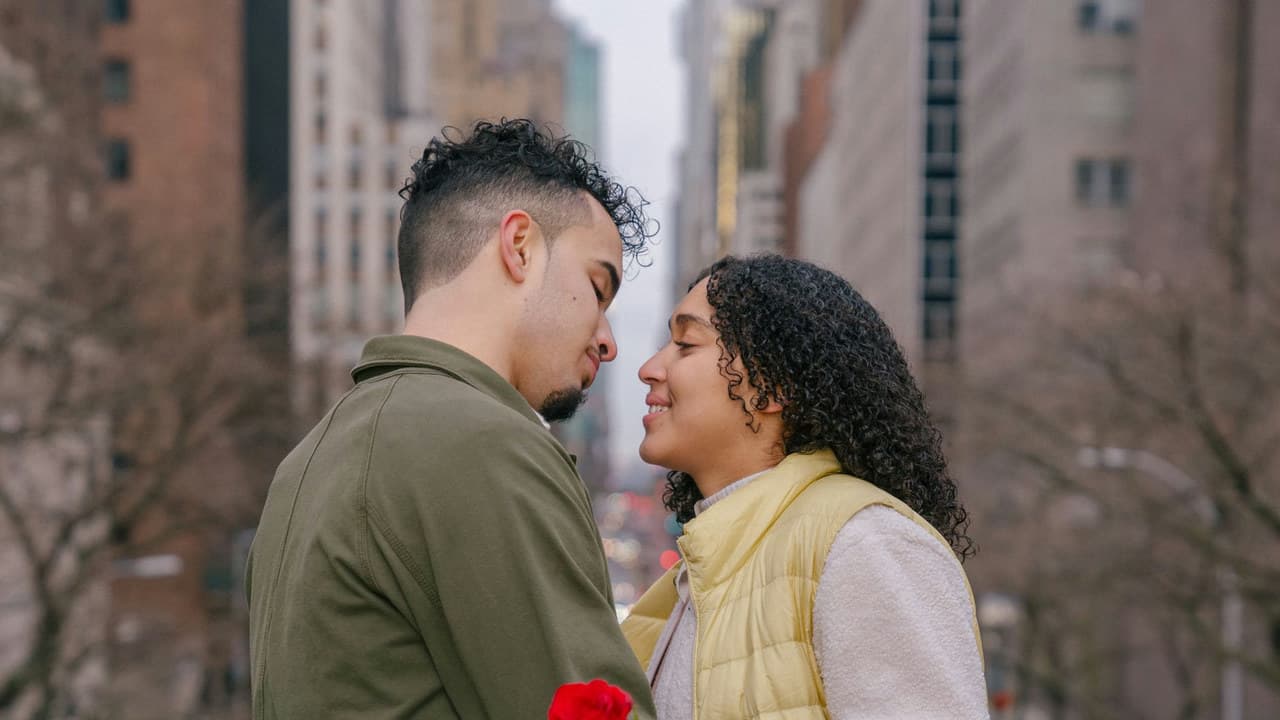 Side view of positive ethnic couple with fresh red rose kissing on blurred background of urban buildings