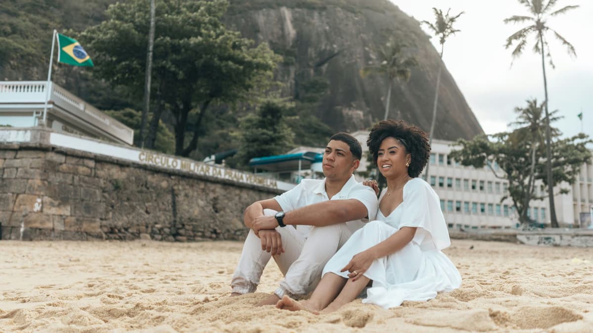 Couple sitting on Copacabana Beach, Brazil, with lush scenery and iconic rock in the background.