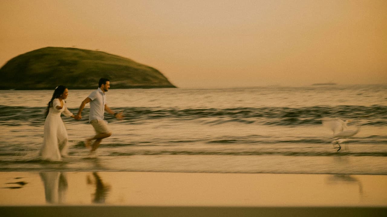 A couple runs joyfully on a beach at sunset with a scenic view of Rio de Janeiro.